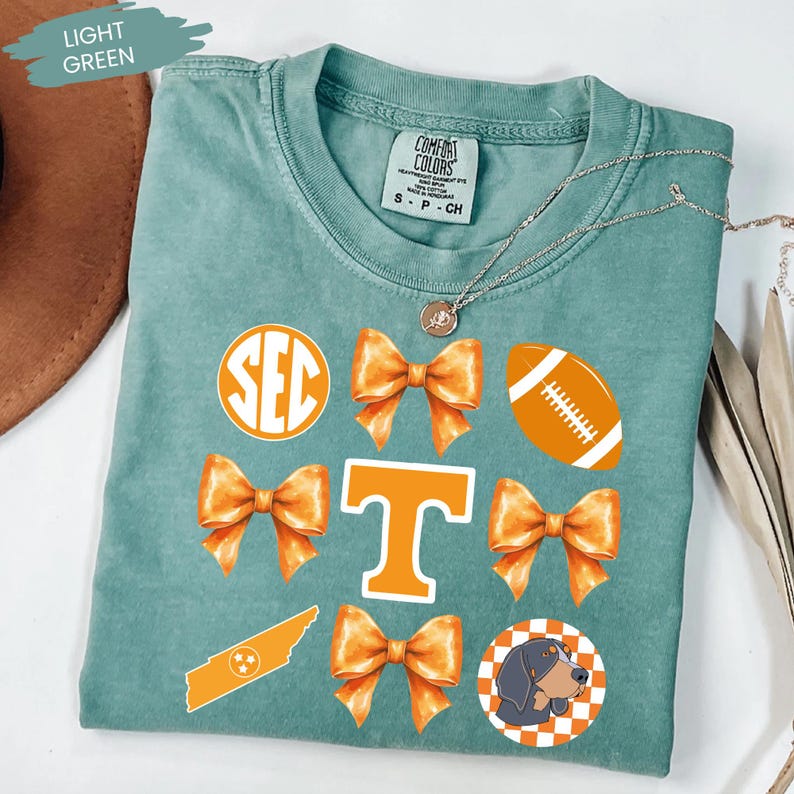 A light green t-shirt with various decorative elements, including a football, a dog, and a ribbon, printed on it.