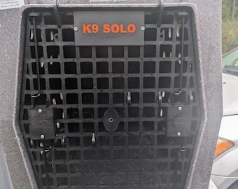 May include: A gray speckled RuffLand Performance Kennels K9 SOLO dog crate featuring a black grid door with two vertical locking latches. The front panel displays the brand name and the model name K9 SOLO in orange text on a gray rectangular plate.