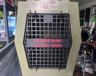 May include: An olive green RuffLand Performance Kennels dog crate featuring a black grid door. A horizontal black sign is attached to the center of the door with the red text FLIGHT RISK DO NOT OPEN printed in a bold, sans-serif font.