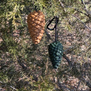 May include: Two pinecone ornaments hanging from a tree branch. One is a light brown color, and the other is dark green. Both are suspended by black cords and carabiner clips.