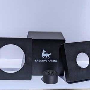 May include: A set of black matte items including two square panels with large circular cutouts, a central square block featuring the white text KREATIVE KANINE with a dog silhouette logo, and a small cylindrical component with multiple holes.