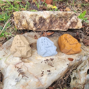 May include: Three decorative rock-shaped objects in beige, light blue, and orange, each with multiple small holes. They rest on a large, light-colored rock, with a larger, rectangular rock above them. The setting is outdoors, with grass and foliage.