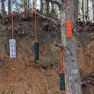 May include: Four cylindrical containers, each suspended from a tree branch by orange rope. The containers are white, black, orange, and green, each with a series of evenly spaced holes. The tree is in a wooded area.