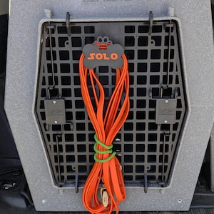 May include: A gray dog crate with a black grid door and an orange leash. The leash is secured with a green rubber band and a bone-shaped tag that reads "SOLO". The crate is labeled "Ruff Land Performance Kennels".