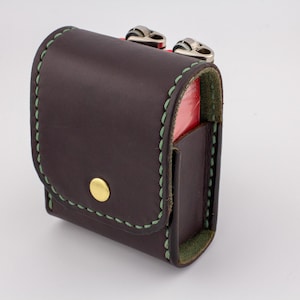 May include: A dark brown leather case with green stitching and a gold button closure. The case has a flap and a small pocket on the side. The interior is red. The case has two silver metal clasps.