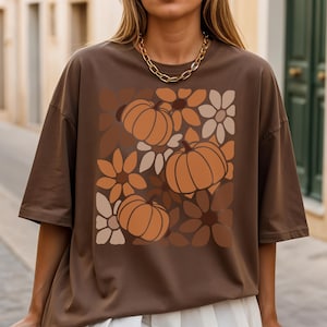 May include: Brown t-shirt with a fall design of pumpkins and flowers in orange, brown, and beige. The oversized tee has short sleeves and a relaxed fit. A great choice for autumn.
