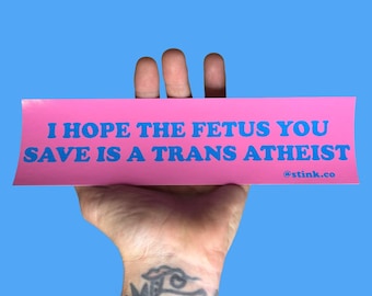 May include: A pink bumper sticker with blue text that reads "I HOPE THE FETUS YOU SAVE IS A TRANS ATHEIST" and the website @stink.co. The sticker is held up against a blue background.