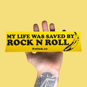May include: A yellow bumper sticker with black text that reads "MY LIFE WAS SAVED BY ROCK N ROLL" and a black banana illustration. The sticker also includes the text "@stink.co".