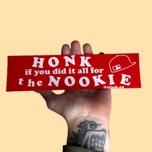 May include: A red bumper sticker with white text that reads "HONK if you did it all for the NOOKIE." A white baseball cap graphic is on the right. The sticker also includes the text "@stink.co."
