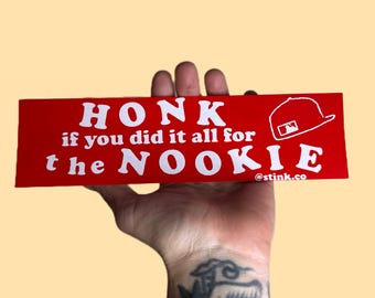 Könnte beinhalten: Roter Aufkleber mit weißem Text: "HONK if you did it all for the NOOKIE." Eine weiße Baseballkappe-Grafik befindet sich auf der rechten Seite. Der Aufkleber enthält auch den Text "@stink.co".