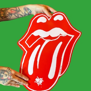 Könnte beinhalten: Ein großes, rotes, stilisiertes Lippenlogo mit weißen Akzenten und einer kleinen Insektengrafik. Das Logo wird vor einem einfarbigen grünen Hintergrund gehalten. Das Logo ist ein erkennbares Design.