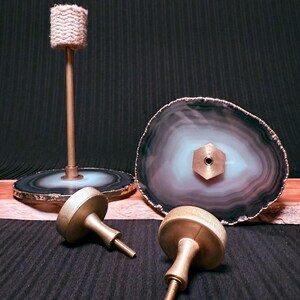 May include: Three gold metal cabinet knobs with a round base and a screw on the bottom. Two of the knobs are lying on a black surface and one is standing upright. Behind the knobs are two agate coasters with a black and white pattern.