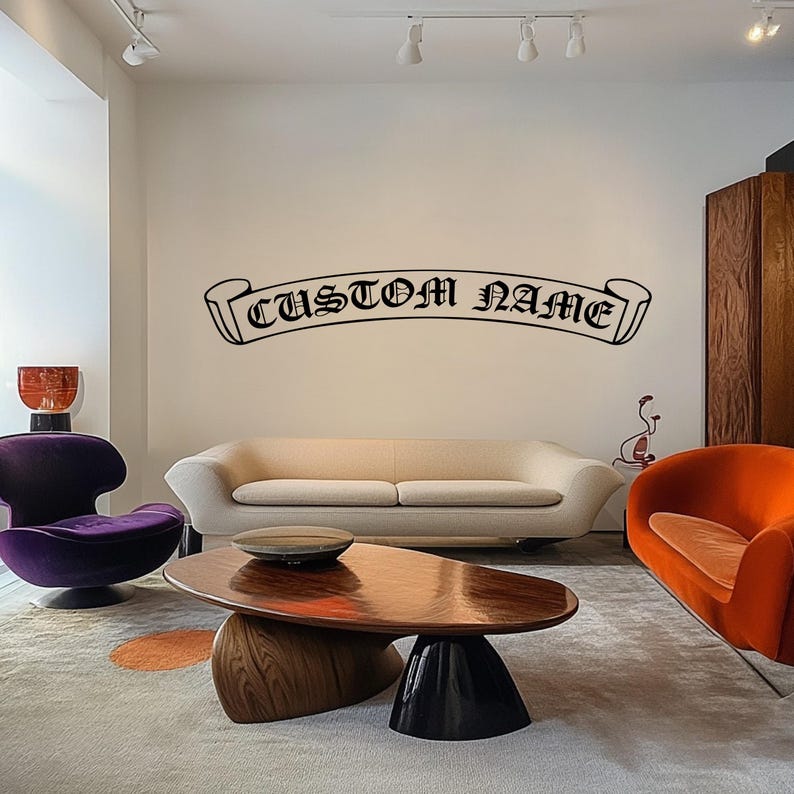 May include: A black and white custom name banner decal with the text "CUSTOM NAME" in a gothic font. The decal is displayed on a white wall above a cream-coloured sofa in a living room.