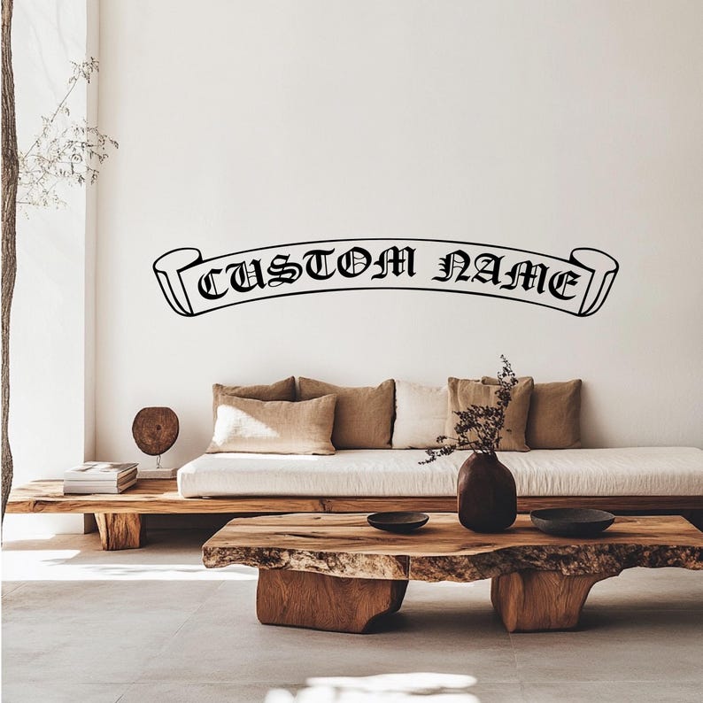 May include: A black and white custom name banner with a scroll design. The banner is on a white wall above a light brown sofa with cushions and a wooden coffee table.