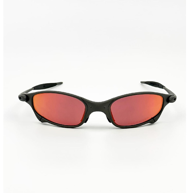 May include: Black sunglasses with red lenses. The sunglasses have a sleek, modern design with a matte black frame. The lenses are a vibrant red colour, offering a bold contrast to the frame. The temples curve slightly for a comfortable fit.