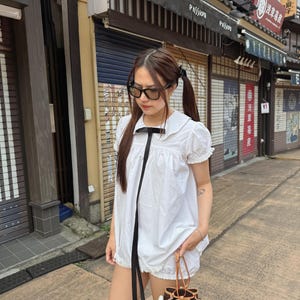 Può includere: Un completo in cotone bianco con colletto Peter Pan e dettagli in nastro nero. Il completo comprende un top e pantaloncini. Una piccola borsa marrone e beige con chiusura a coulisse è tenuta in mano, insieme a un piccolo peluche.