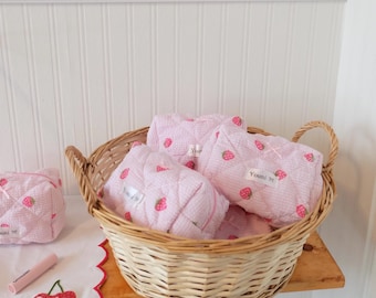 May include: A wicker basket filled with pink quilted pouches with strawberry patterns. The pouches have the text "Youmi 98" printed on them. The basket is sitting on a wooden shelf with a white wainscoting wall behind it.