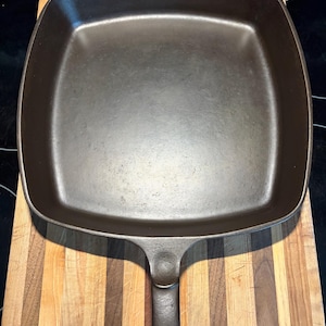 May include: A square, cast iron skillet with a long handle. The pan is dark gray and sits on a wooden cutting board with vertical stripes of light and dark wood. The skillet is empty and ready for cooking.