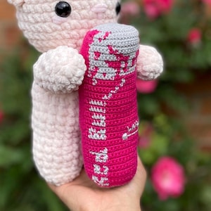 May include: A crocheted pink bunny holding a pink and grey drinks can cosy. The bunny is light pink with black eyes. The can cosy has white text and a grey top. The cosy is wrapped around a drinks can.