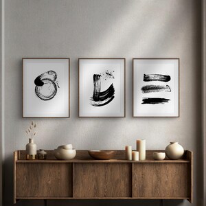 Peut inclure: Trois impressions d'art abstrait noir et blanc dans des cadres simples, accrochés à un mur gris clair au-dessus d'un meuble en bois avec un bol et des bougies sur le dessus.