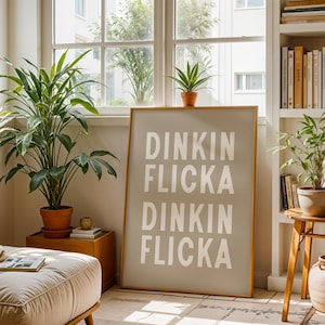 May include: A framed print with the words "Dinkin Flicka" in large white letters on a light grey background. The print is in a room with plants, a window, and a wooden side table.