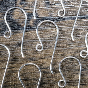 Op de afbeelding: Close-up van verschillende zilverkleurige oorhaakjes. De haakjes hebben een gebogen ontwerp met een lusvormig uiteinde en een rechte pin. Ze zijn gerangschikt op een getextureerd, donkerbruin houten oppervlak, waardoor een patroon ontstaat.