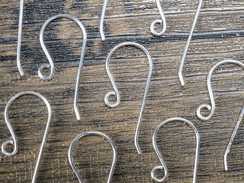 K&ouml;nnte beinhalten: Nahaufnahme von mehreren silberfarbenen Ohrhaken. Die Haken haben ein gebogenes Design mit einem geschlungenen Ende und einem geraden Stift. Sie sind auf einer strukturierten, dunkelbraunen Holzoberfl&auml;che angeordnet und bilden ein Muster.