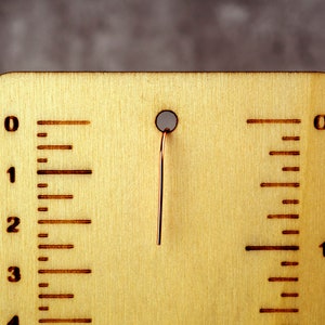 May include: A wooden ruler with etched markings and a copper wire. The ruler is light brown with dark brown markings indicating inches. The wire extends from a hole at the top. The ruler measures up to 4 inches.