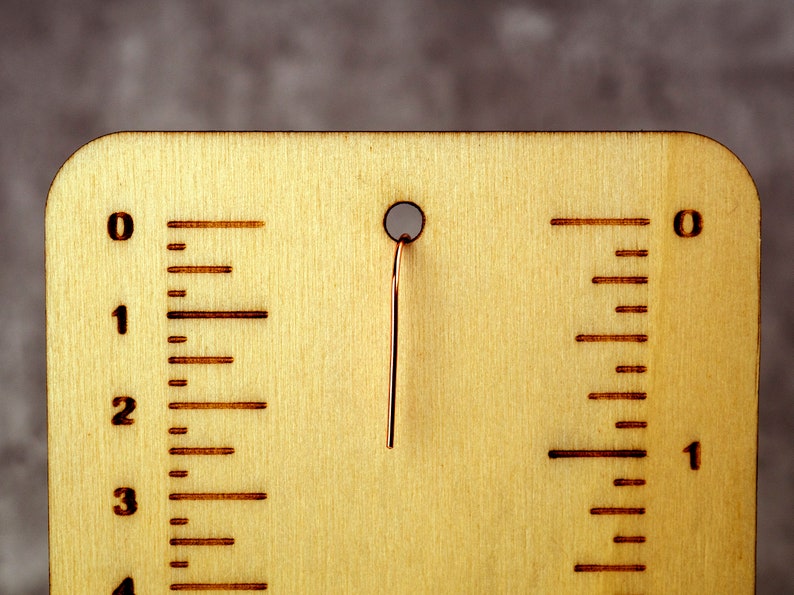 May include: A wooden ruler with etched markings and a copper wire. The ruler is light brown with dark brown markings indicating inches. The wire extends from a hole at the top. The ruler measures up to 4 inches.