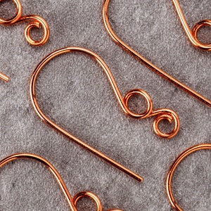May include: Close-up of several copper-colored earring hooks. The hooks have a simple, curved design with a looped end. The background is a textured gray fabric. The earrings are likely handmade.