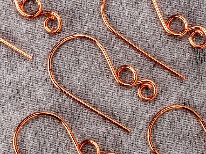 May include: Close-up of several copper-colored earring hooks. The hooks have a simple, curved design with a looped end. The background is a textured gray fabric. The earrings are likely handmade.