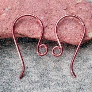 May include: Two copper-colored earring hooks are displayed against a textured, pinkish-red stone. The hooks have a simple, curved design with a small, circular detail at the bottom. The background is a gray, patterned fabric.