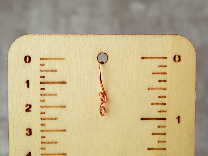 May include: A wooden measuring tool with etched markings and a copper wire accent. The tool features inch measurements, with numbers and lines indicating increments. A small hole is at the top, with a decorative copper wire hanging from it.