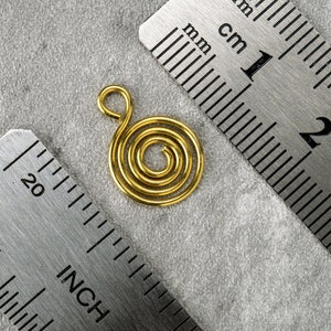 Può includere: Un ciondolo a spirale color oro con un anello nella parte superiore. Il ciondolo ha un diametro di circa 2,5 cm. Un righello di metallo con misure in cm è in primo piano.