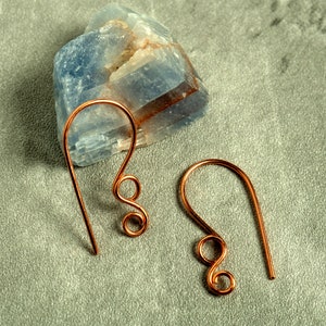 May include: A pair of copper-colored wire earrings with a hook design and a decorative swirl. The earrings are displayed on a gray surface with a blue and white stone in the background.