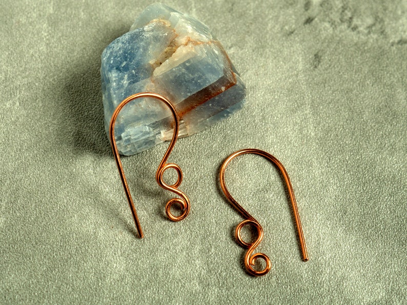 May include: A pair of copper-colored wire earrings with a hook design and a decorative swirl. The earrings are displayed on a gray surface with a blue and white stone in the background.