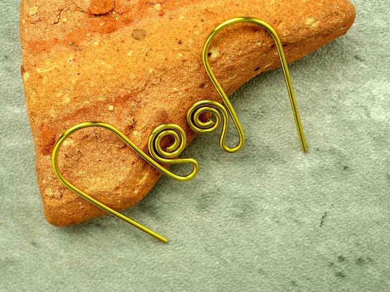 A pair of brass French hook ear wires featuring a small spiral are shown propped against a chunk of terracotta. The ear wires are compact and chunky looking. The background is a mottled grey.