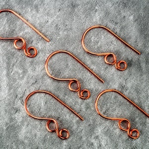 May include: Five copper-colored earring hooks are arranged on a gray surface. Each hook has a curved top and a straight, pointed bottom. The design includes a decorative swirl near the bottom, suggesting a handmade jewelry component.