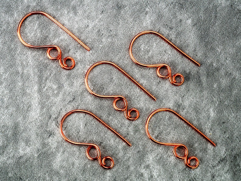 May include: Five copper-colored earring hooks are arranged on a gray surface. Each hook has a curved top and a straight, pointed bottom. The design includes a decorative swirl near the bottom, suggesting a handmade jewelry component.