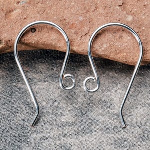 Op de afbeelding: Een paar zilverkleurige, gebogen oorhaken. Elke haak heeft een lusvormig ontwerp aan de bovenkant en een gebogen onderkant. De oorbellen worden getoond op een getextureerd, grijs oppervlak met een roodbruine steen op de achtergrond.