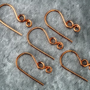 May include: Five copper-colored earring hooks are arranged on a gray, textured surface. Each hook has a curved design with a looped end. The hooks are made of thin wire and are evenly spaced.