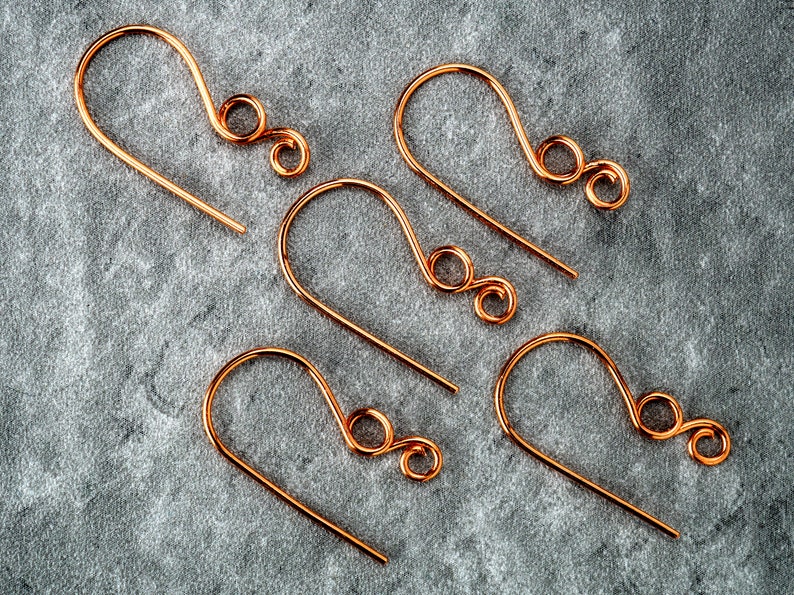 May include: Five copper-colored earring hooks are arranged on a gray, textured surface. Each hook has a curved design with a looped end. The hooks are made of thin wire and are evenly spaced.