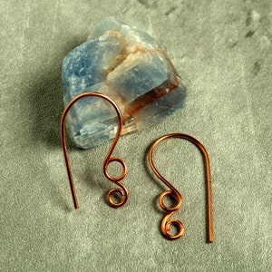 May include: A pair of copper-coloured wire earrings with a looped design. The earrings are displayed on a grey surface next to a blue and white crystal. The earrings have a simple, elegant design.