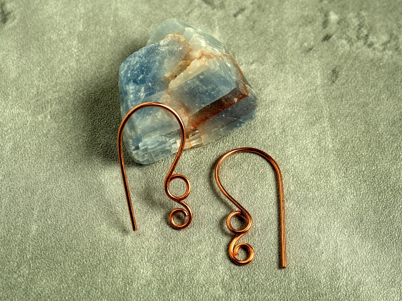 May include: A pair of copper-colored wire earrings with a looped design. The earrings are displayed on a gray surface next to a blue and white crystal. The earrings have a simple, elegant design.