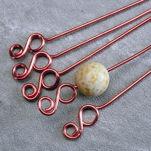 May include: Four copper-colored metal skewers with decorative swirl ends. One skewer has a speckled, round, off-white bead. The skewers are arranged diagonally against a gray textured background. The design is simple and elegant.