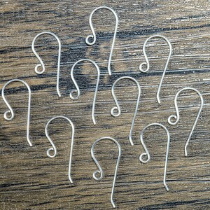 K&ouml;nnte beinhalten: Eine Ansammlung silberfarbener Ohrhaken, angeordnet auf einer Holzoberfl&auml;che. Jeder Haken hat ein gebogenes Design mit einem geschlungenen Ende. Die Haken sind gleichm&auml;&szlig;ig verteilt und zeigen ihr schlichtes, aber elegantes Design. Geeignet f&uuml;r die Schmuckherstellung.