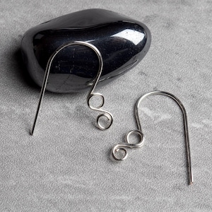 May include: A pair of silver-colored, wire-wrapped earrings. Each earring features a curved hook design with a decorative swirl at the bottom. The earrings are displayed on a gray surface with a dark, polished stone in the background.