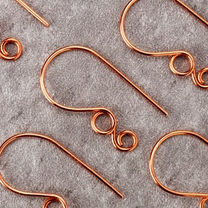 May include: Close-up of several copper-colored earring hooks. Each hook has a curved design with a looped end and a straight wire for insertion. The hooks are arranged on a gray fabric surface, showcasing their intricate design.