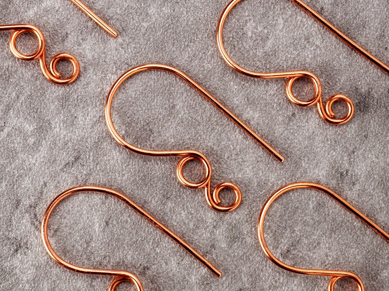 May include: Close-up of several copper-colored earring hooks. Each hook has a curved design with a looped end and a straight wire for insertion. The hooks are arranged on a gray fabric surface, showcasing their intricate design.