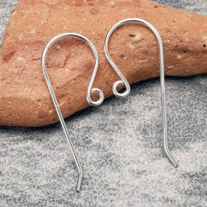 K&ouml;nnte beinhalten: Zwei silberfarbene Ohrhaken. Jeder Haken hat eine gebogene Oberseite mit einer kleinen, aufgerollten Schlaufe und einen geraden, schlanken Draht, der sich am Ende kr&uuml;mmt. Die Haken werden auf einem strukturierten, rotbraunen Stein vor grauem Hintergrund gezeigt.